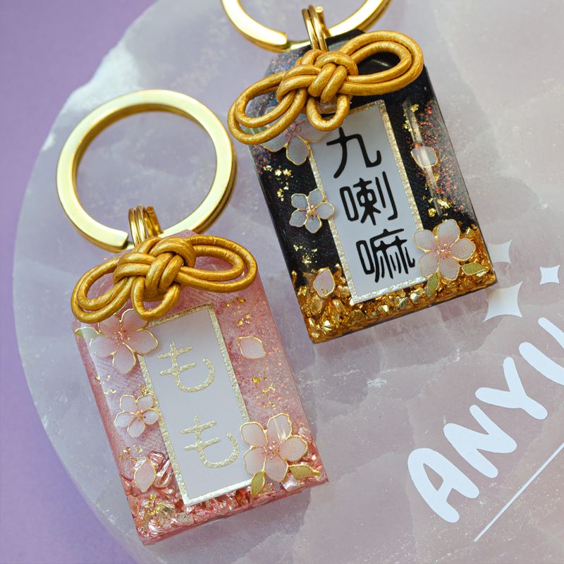 A picture of Hundemarke in Omamori-Form. Das Design besteht aus fallenden Kirschblüten mit Akzenten aus Blattgold und in der Marke sind Kristallsteine enthalten. Die Hundemarke wird außerdem mit einem handgebundenen Knoten versehen.